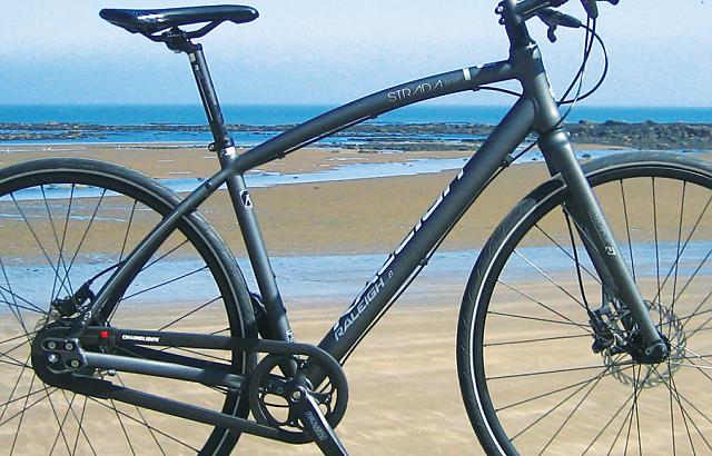 A dark grey hybrid bike with flat handlebar, disc brakes and no mudguards. In the background is a beach with the tide far out