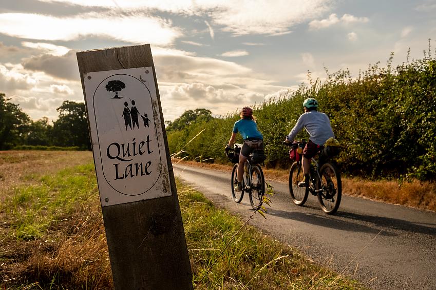 Two cyclist ride along a lane by a hedgerow with a sign in the foreground saying "Quiet lane"