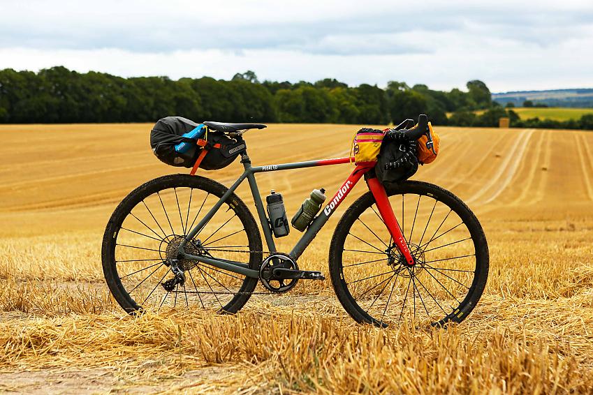 A grey and red gravel bike with packed bar and saddle bags is propped up in a mown field of hay with a line of trees in the background