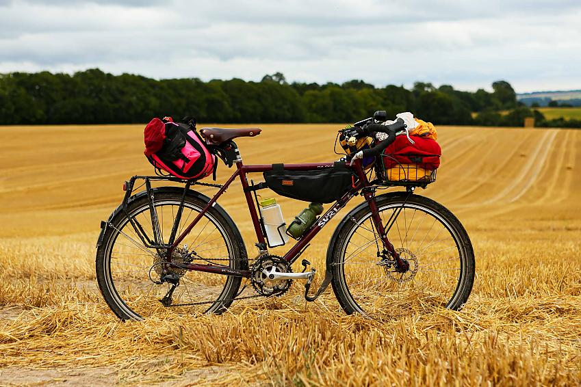 A maroon touring bike complete with packed front rack, saddle bag and bar bag is propped up in a mown field of hay with trees in the background