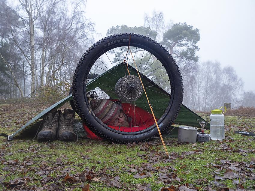 A man sheltering under a tarp using a bicycle wheel to keep it up
