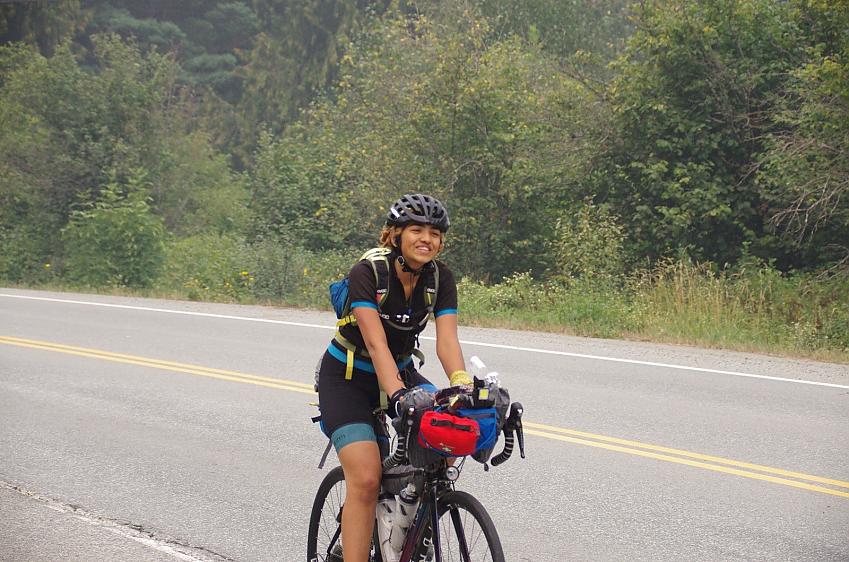A woman cyclist is cycle touring. She is riding along a road with trees in the background. Her bike is packed for a long trip.