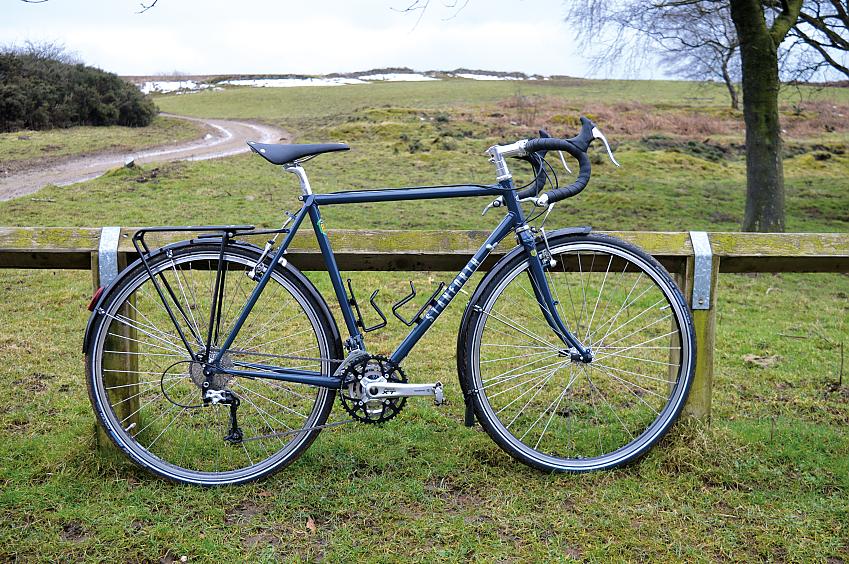 Stanforth Skyelander, a dark blue touring bike leaning against a wooden fence. It's got a drop handlebar, rear luggage rack and two bottle cages