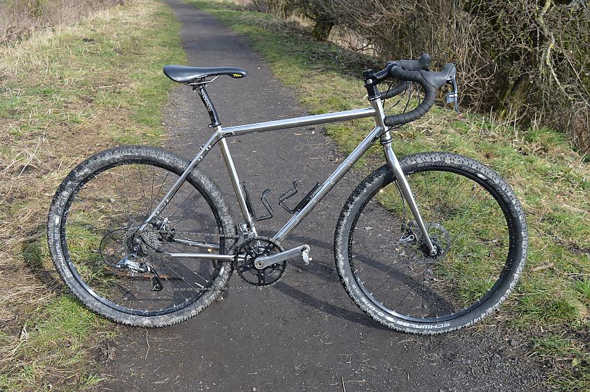 A silver touring bike propped up in a country lane. It's got drop bars, fat tyres and no rack or frame bags