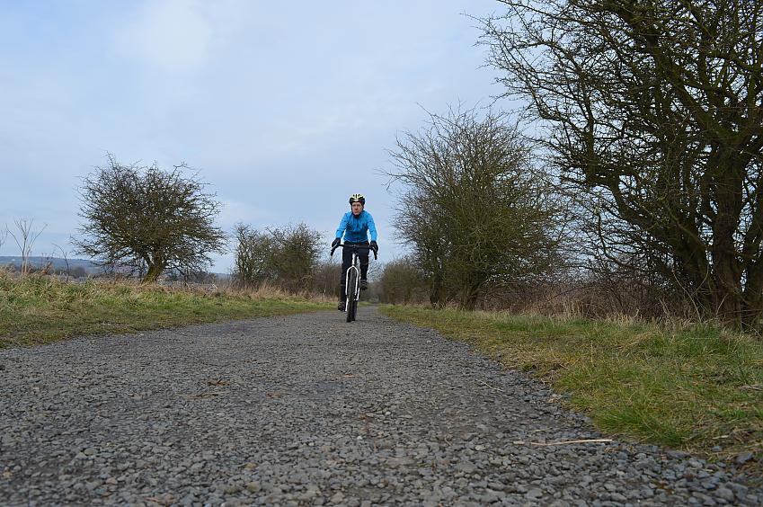 A man is cycling along a gravel path. He's wearing black cycling trousers and a blue waterproof jacket; it looks quite overcast