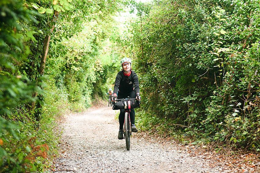 A woman is cycling along an off-road gravel path. Both sides of the path are lined with trees and hedges and lots of greenery.