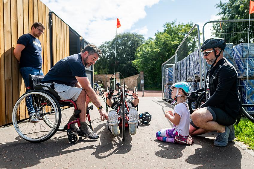 A wheelchair user helps a handcyclist get set up as other cyclists watch on