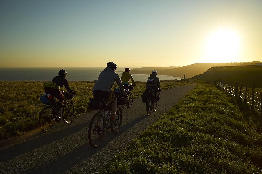 Four cyclists are riding away from the camera along a clifftop path. The sea is to their left and the sun is setting ahead