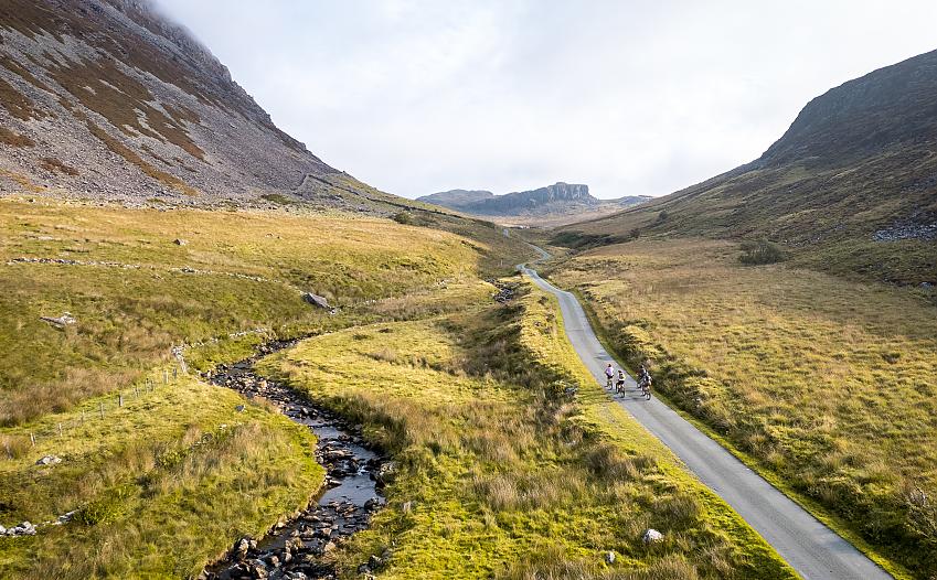 An aerial image of four cyclists riding away from the camera on a narrow road in an epic valley. There is a stream next to the road and mountains on both sides and up ahead.