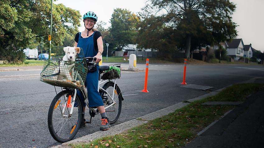 Woman stands with her bike with a dog in the basket in a cycle lane marked with traffic wands