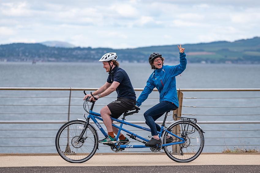 Two people riding a tandem past a loch