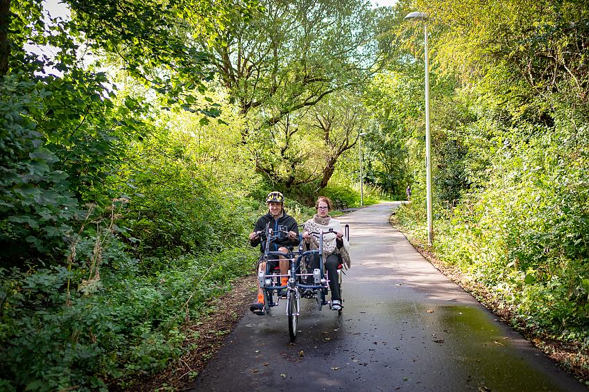 Two people riding an adapted trike along a cycle path through trees