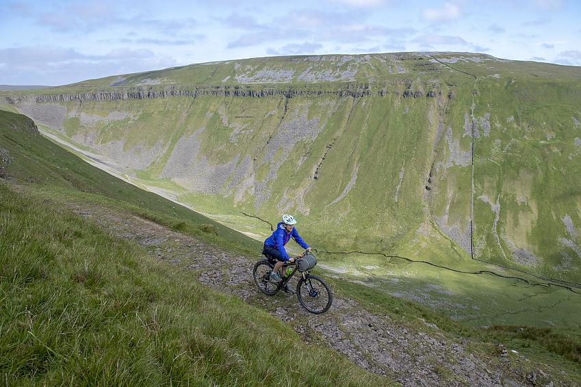 A mountain biker rides along a stony trail along the side of a dramatic valley at High Cup Nick, Cumbria