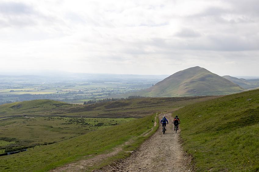 Two mountain bikers riding down a gravel track towards hills in the distance.