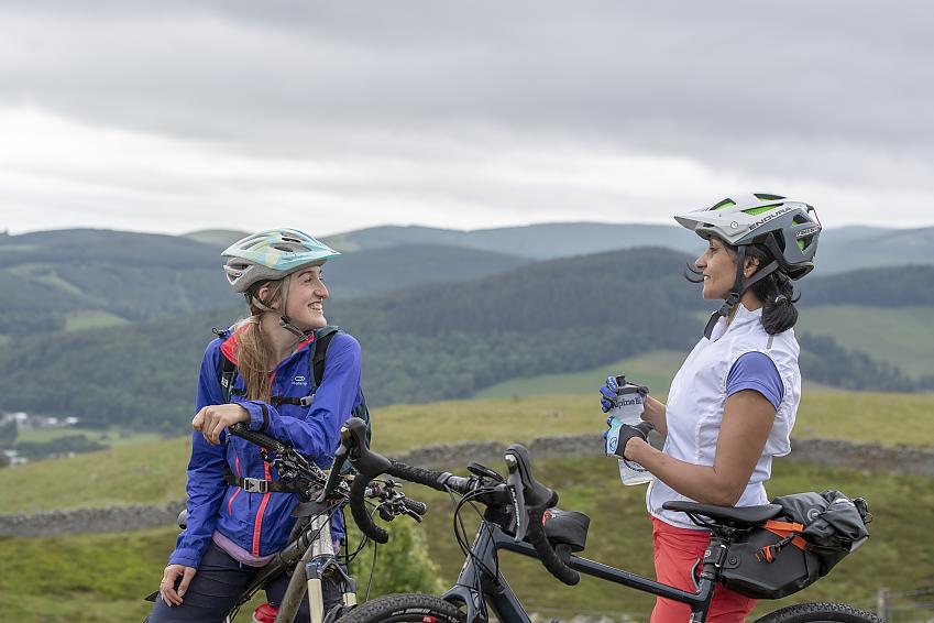 Two women standing with their bikes stop to chat, in front of a view of rolling green hills