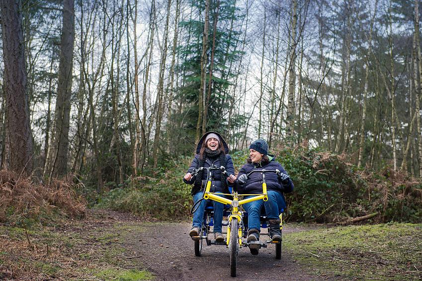 Two women riding together on an adapted tricycle along a woodland trail