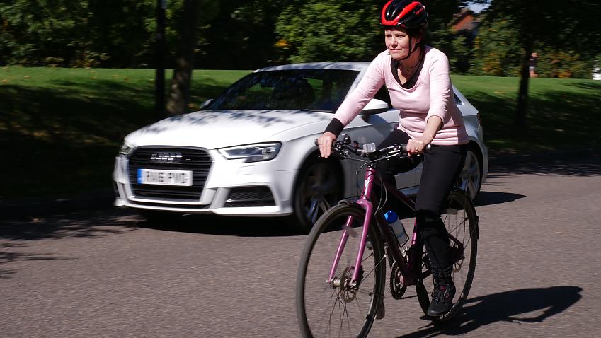 A person driving a white car overtakes a woman cycling