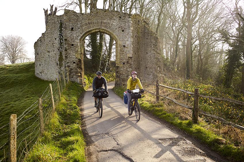 Two people cycle along a country lane beneath a medieval archway