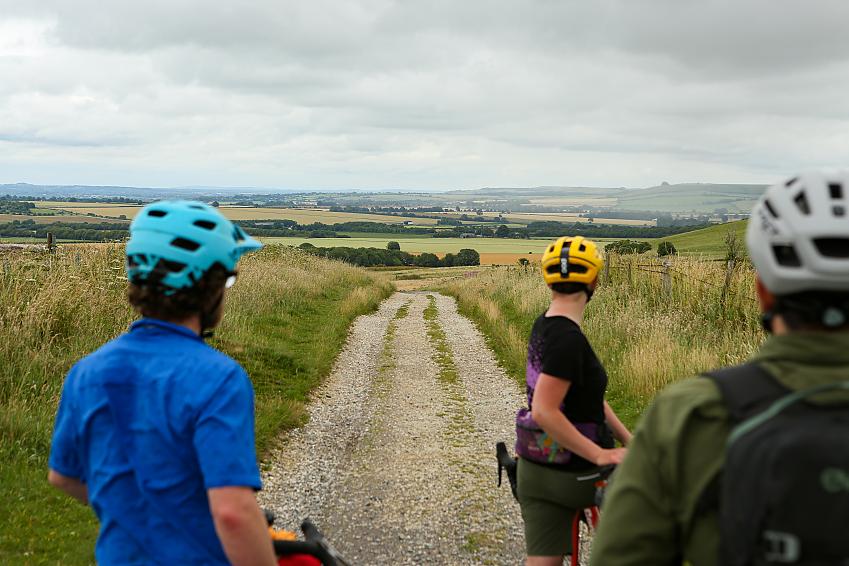 People wearing cycle helmets look down a gravel track that stretches to the hills in the distance