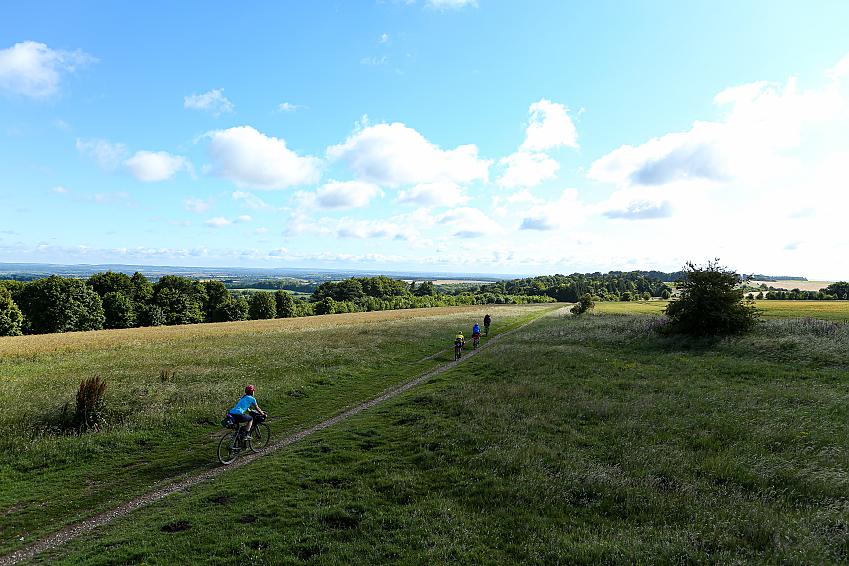 Four people cycle along a grassy track under a wide blue sky