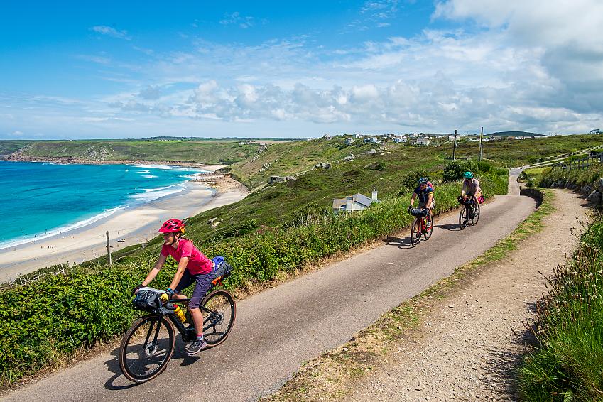 Four people cycle along a road with a stunning beach in the background below