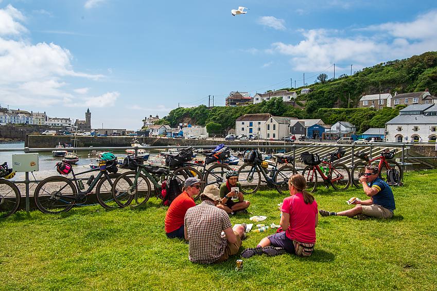 A group of people sit eating pasties beside a harbour with their bikes leant against the railings