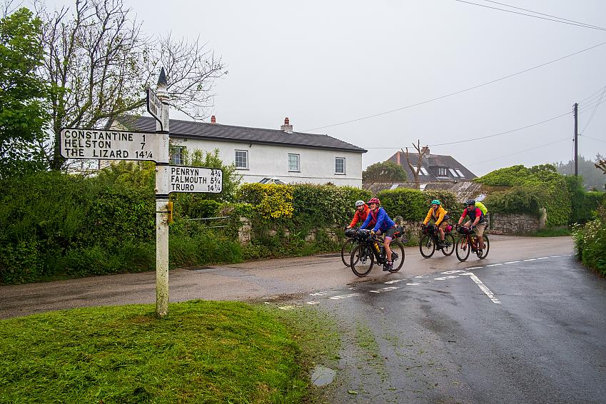 Four people cycle past a signpost on a rural lane