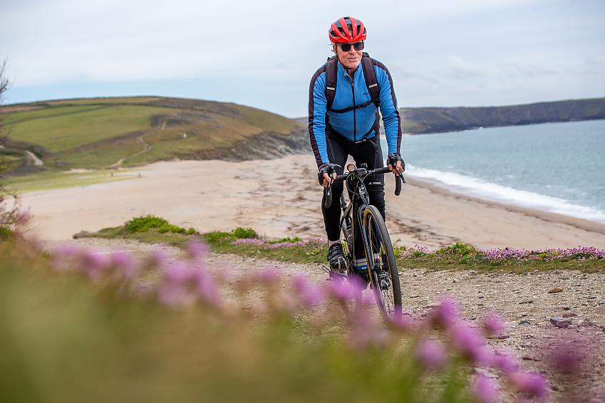 Through pink flowers in the foreground, a man cycles up a hill with a beautiful beach behind
