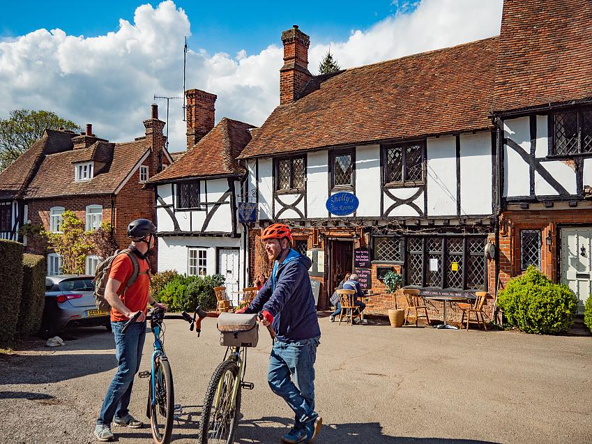 Two men stand with bikes chatting in front of a picturesque Tudor cottage