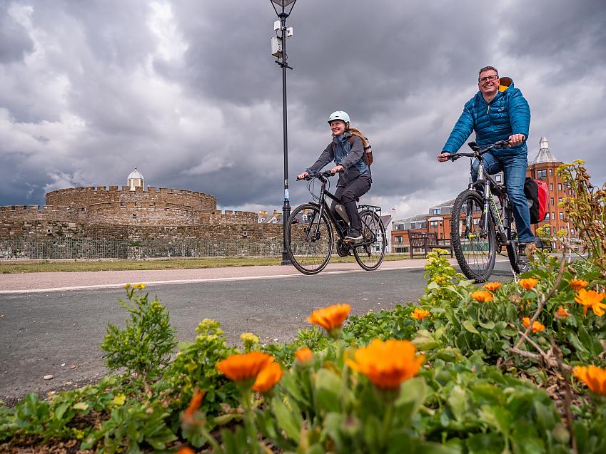 Two people cycle past a castle with orange flowers in the foreground