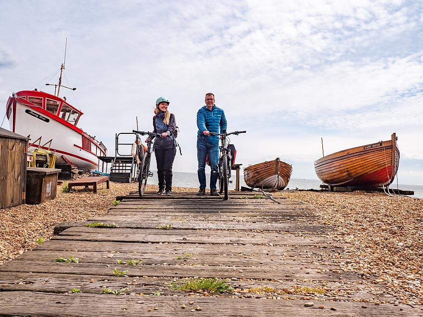 Two people wheeling bikes along a boardwalk on a shingle beach past brightly coloured boats