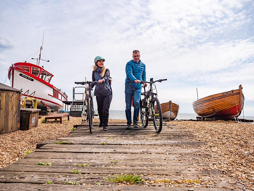Two people push their bikes on a walkway across a shingle beach past colourful boats