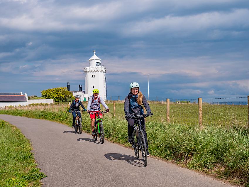People cycle past a white lighthouse in the sunshine