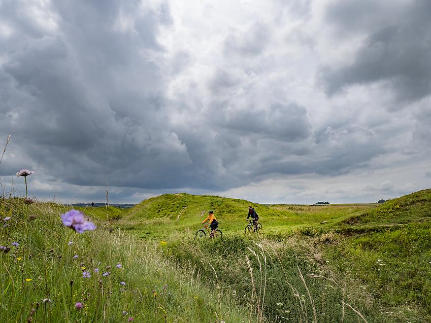 Two people cycle past the grassy mound of Barbury Castle hill fort in Wiltshire