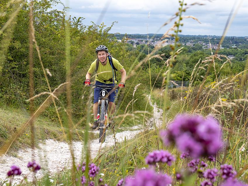 Through a foreground of bright pink flowers, a man on a mountain bike rides up a hill towards the camera