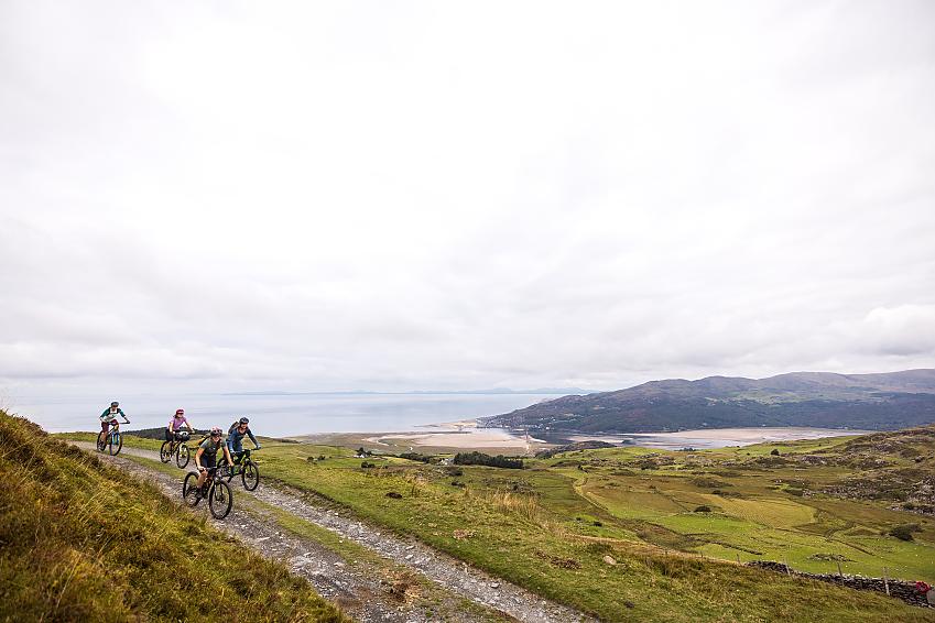 Four women ride mountain bikes on a gravel track high above an estuary