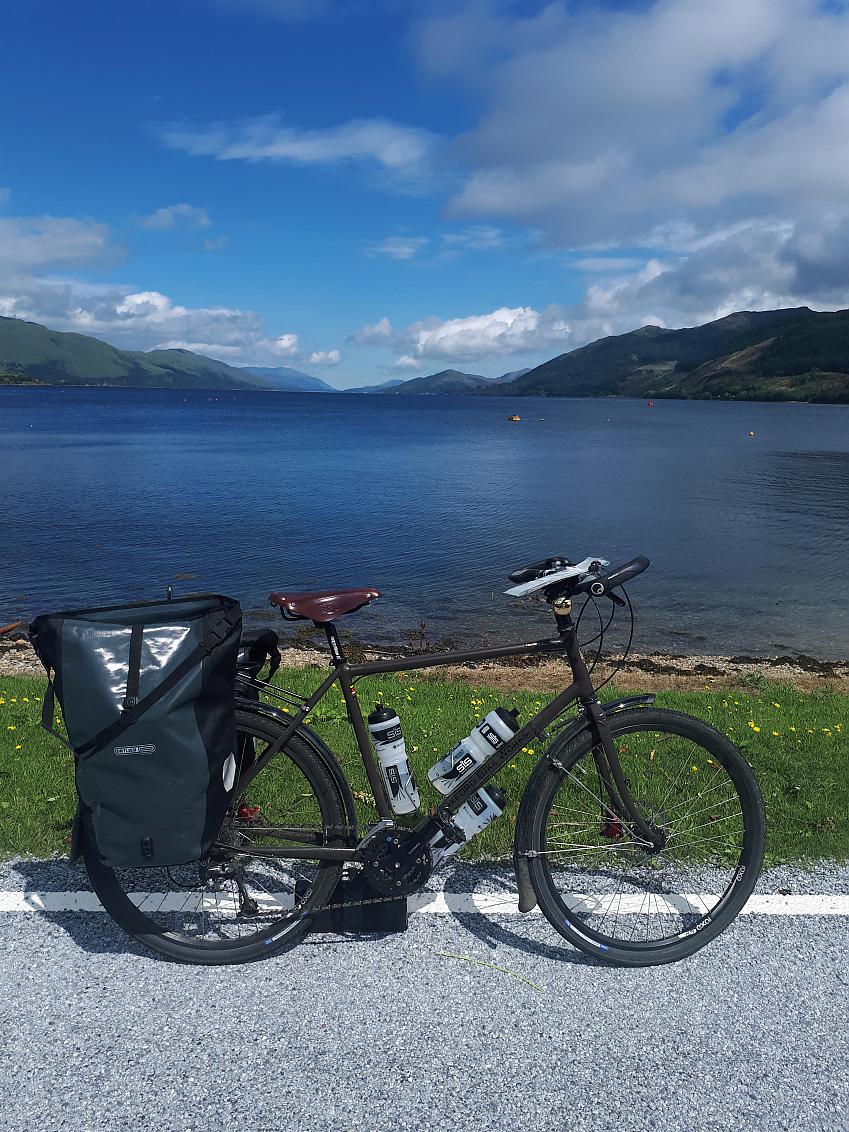 An Oxford Bike Works tourer is upright on a road in front of a large lake surrounded by mountains