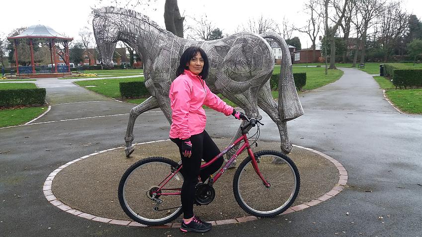 Intensive care nurse, Narinder Kaur. She is standing with her pink mountain bike in front of a metal statue of a horse. It's a wet day and she's wearing a pink waterproof coat and black leggings.