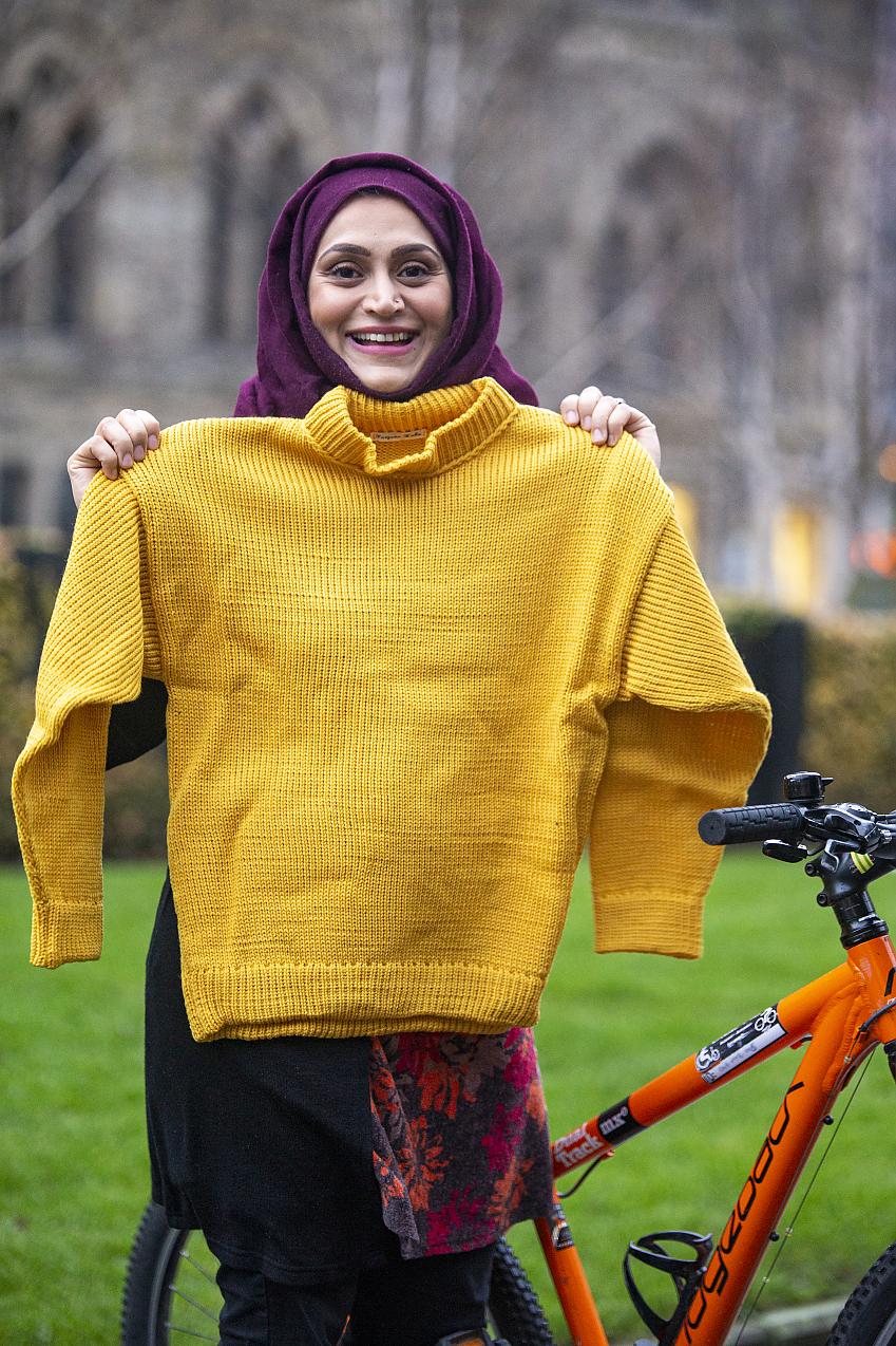 A woman in a purple hijab holds up a knitted yellow sweater 