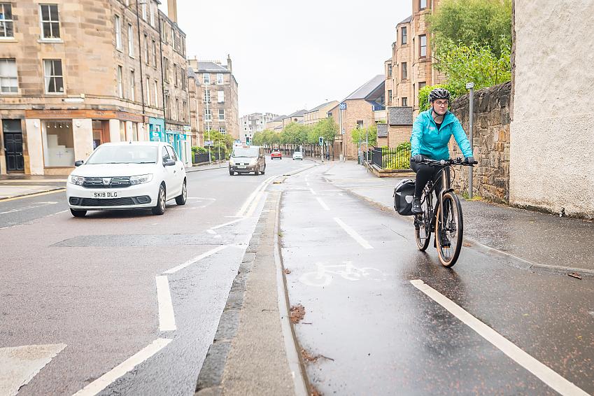 A stretch of segregated cycle path in Edinburgh, with a cyclist cycling along in it. It's raining and the cyclist is wearing a turquoise waterproof jacket. Cars are driving alongside.