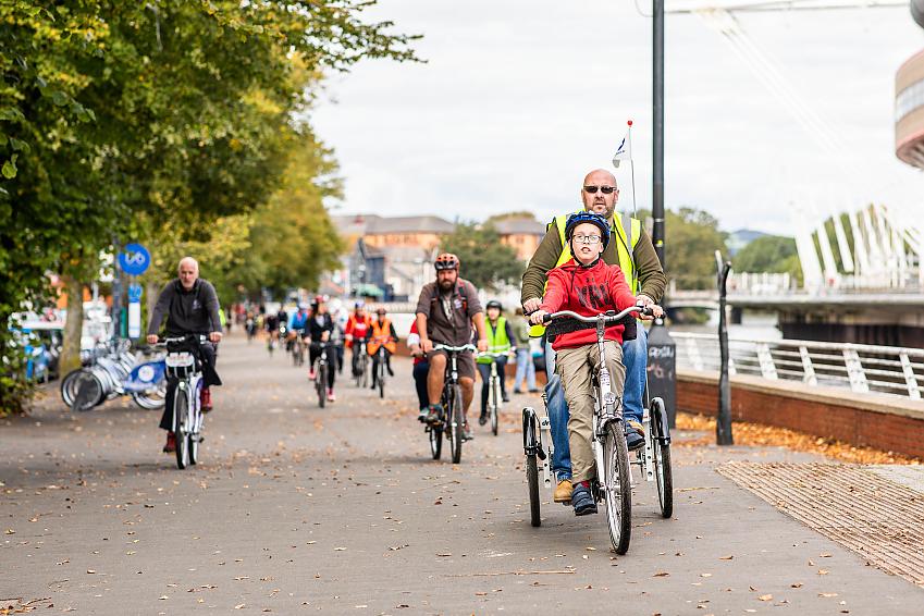 A large number of people are cycling along a cycle path next to a river. At the front is a young boy cycling on a trike with an older man
