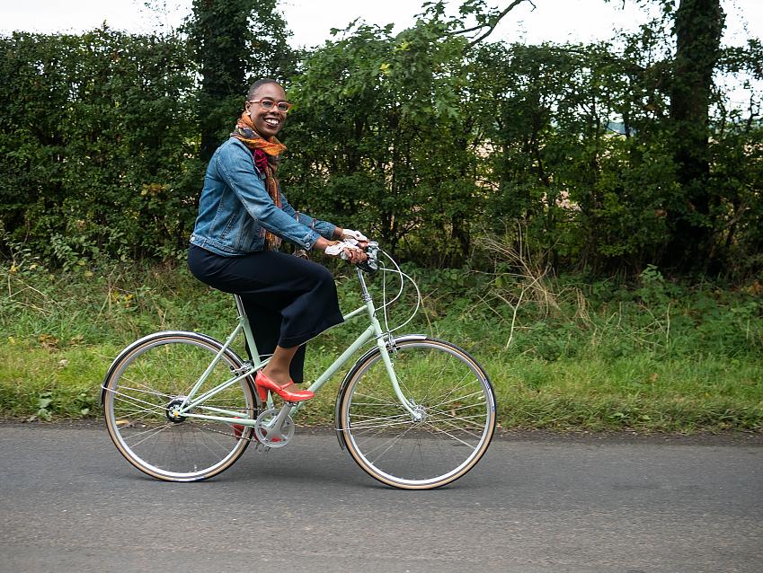 A woman cycling. She is wearing normal clothes – a denim jacket, navy culottes and red high heels. She is smiling.