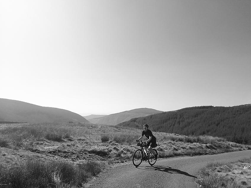 A black and white shot of a woman on a bike as she cycles through Welsh hills
