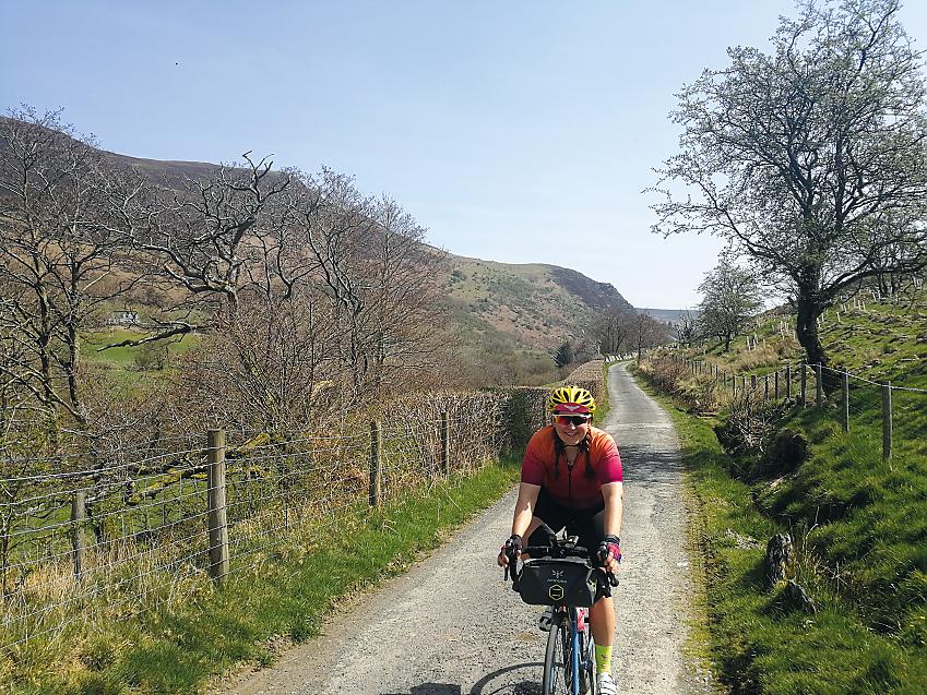 A woman on a blue bike. She is wearing black cycling shorts and a red and orange jersey, as well as a bright yellow helmet. She is smiling as she rides through the Wye valley