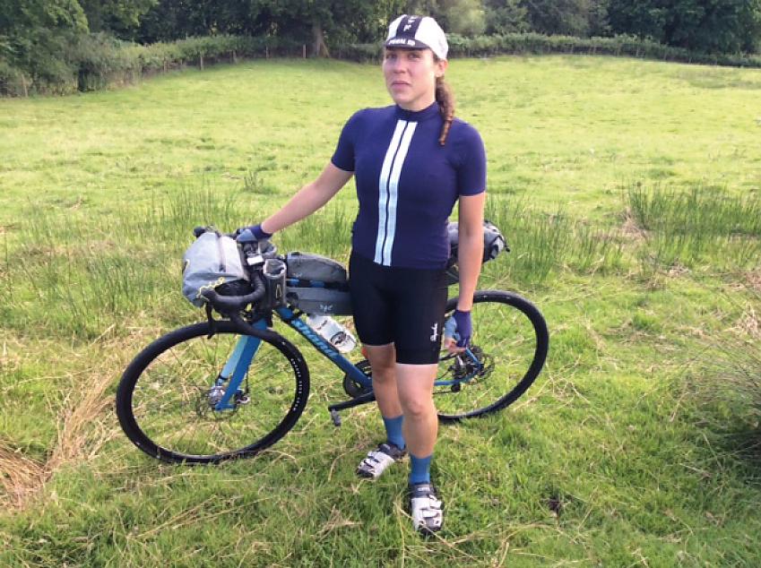 A woman poses with her bike. She is wearing a blue cycling jersey with two vertical white stripes down the front. Her bike is blue too. 