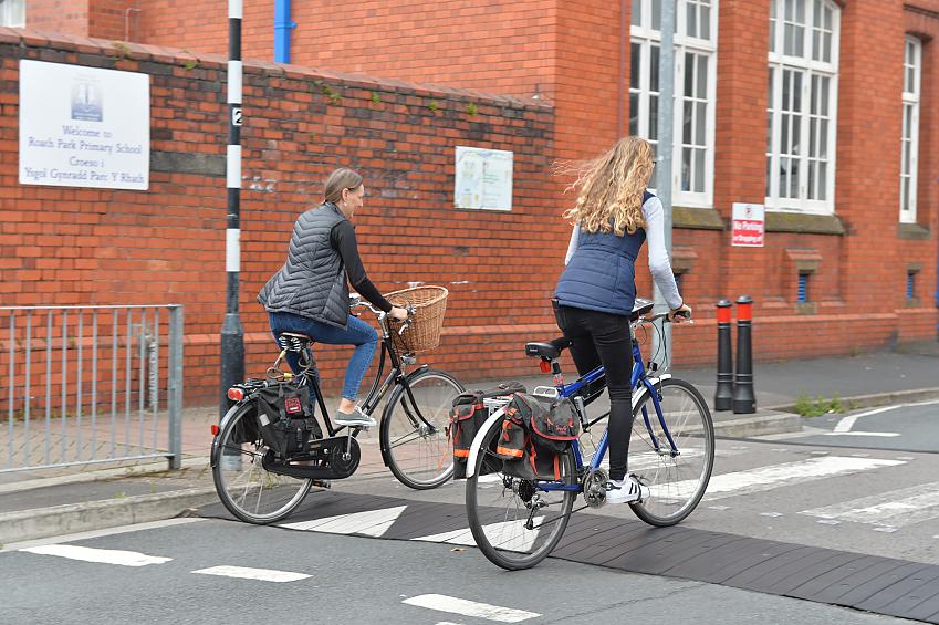 Two women cycle along a road outside school gates. They're crossing a zebra crossing. They're wearing casual clothes, their pannier bags loaded onto the back of their bikes. They are both casually dressed without helmets