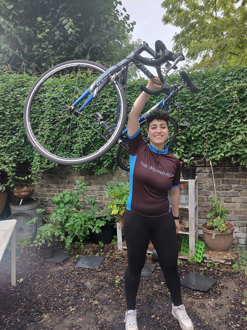 A woman is standing in a garden. She's holding a blue Trek racing bike over her head. She's wearing black leggings and a blue and brown Cycle Project cycling jersey.