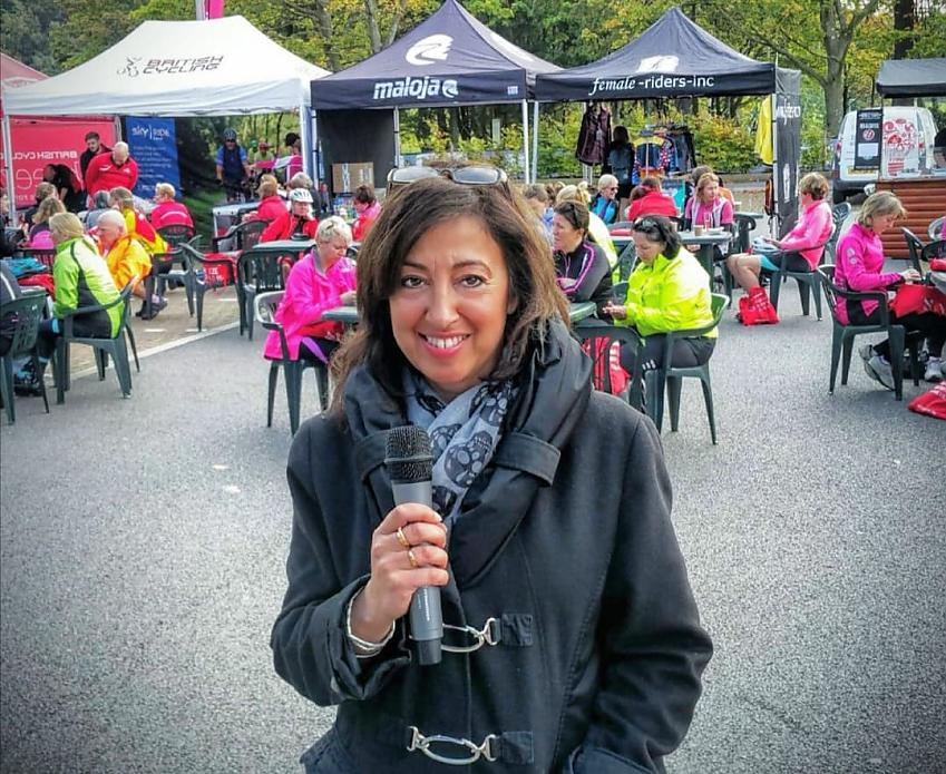 A woman with a microphone is reporting from a cycling event. She's smiling and wearing a grey jacket. Behind her are lots of female cyclists sitting at tables, as well as some stands run by cycling organisations such as British Cycling and Maloja