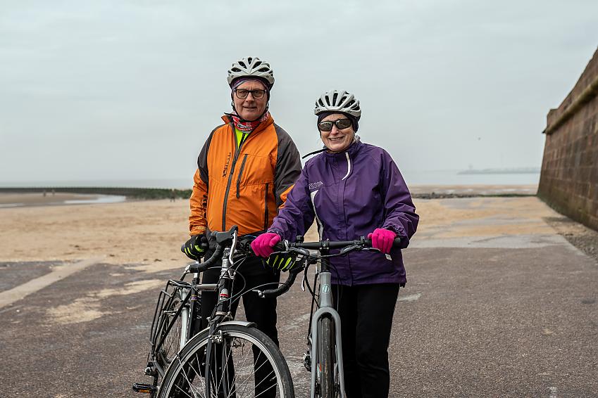 Two people are standing on a beach, holding their bikes. It looks like it's winter, with both wearing waterproof jackets. The man has an orange jacket and a black touring bike; the woman's jacket is purple and she has a grey hybrid bike