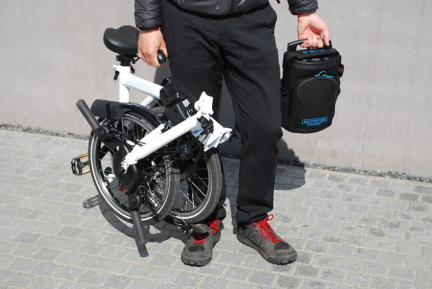 A man is carrying a folded Brompton e-bike in one hand with the bike's battery in the other